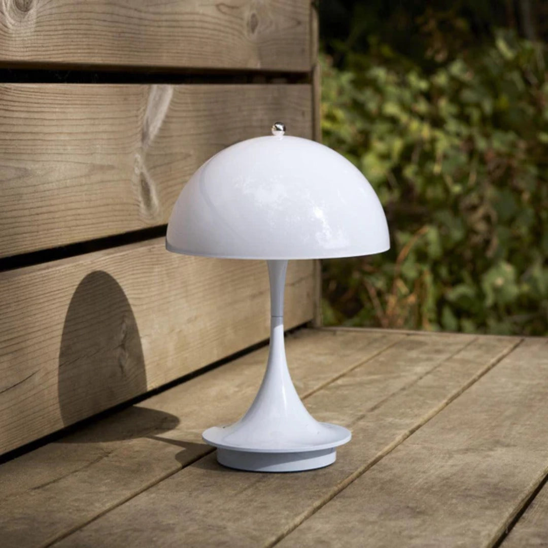 Adjustable gold dome desk lamp with reflective brass finish featuring a white shade, situated on a wooden rectangular table beside a green plant, on hardwood flooring.