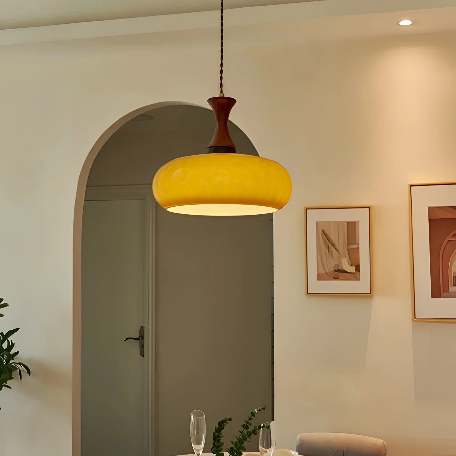Amber glass retro pendant light with a mid-century modern design hanging from the ceiling in a stylish interior featuring wood accents, indoor plants, picture frames, and natural lighting through a window.