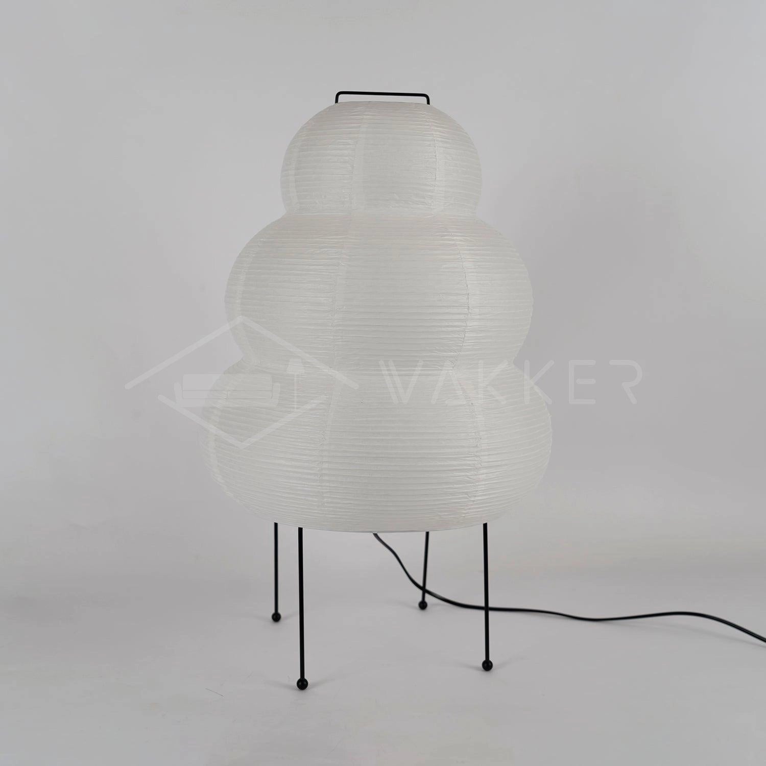 A modern tripod floor lamp with a sculptural beehive design, positioned next to a table and chair in a room with grey tones.