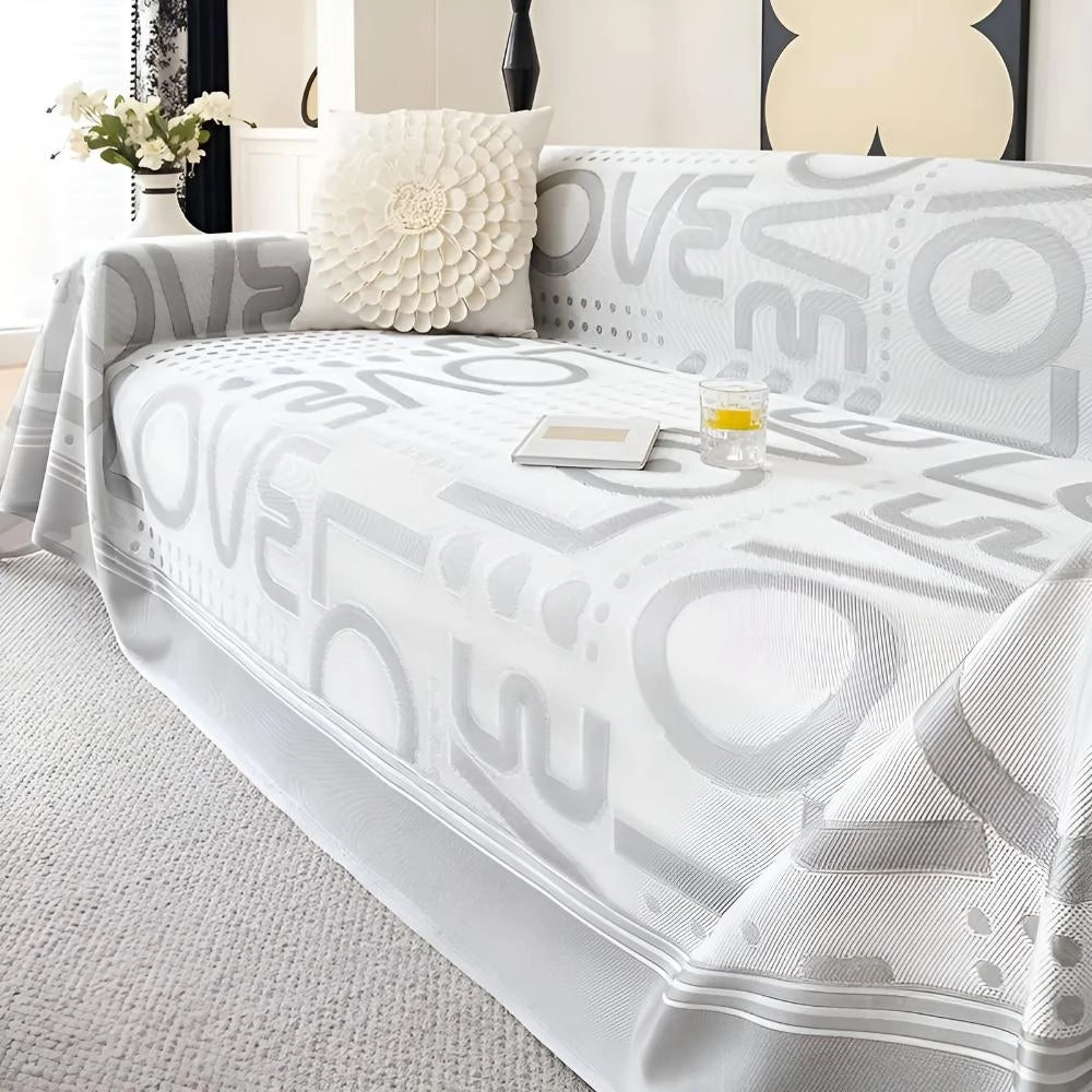 A beige geometric sofa cover elegantly draped over a large grey sofa, measuring 180x420cm, enhancing modern decor with stylish accessories. The scene includes comfortable pillows and a nearby green plant adding a touch of nature.