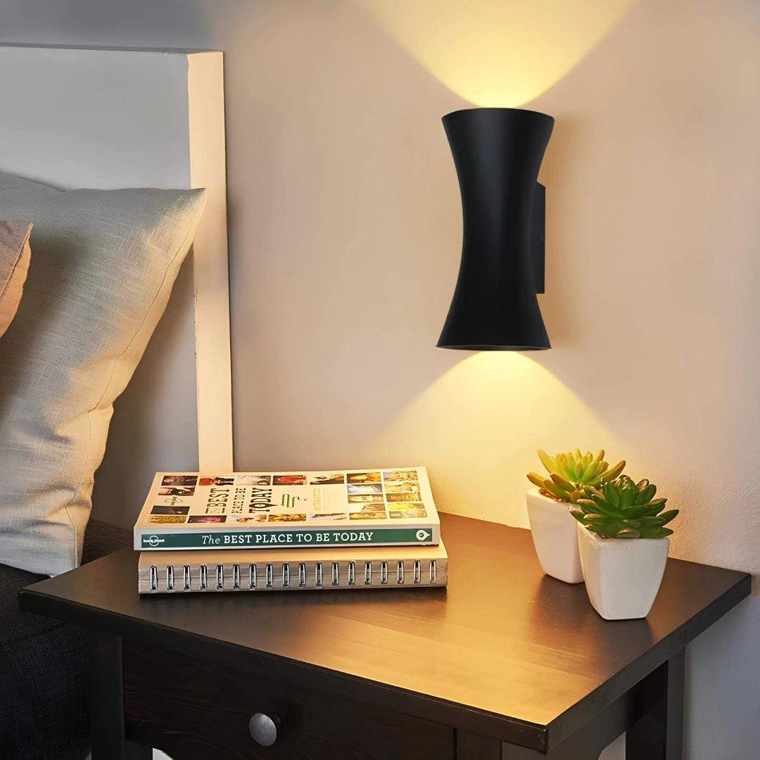 Black outdoor LED wall sconce mounted on an exterior wall, emitting light both upwards and downwards, surrounded by plants and wooden furniture, showcasing its weather-resistant design.