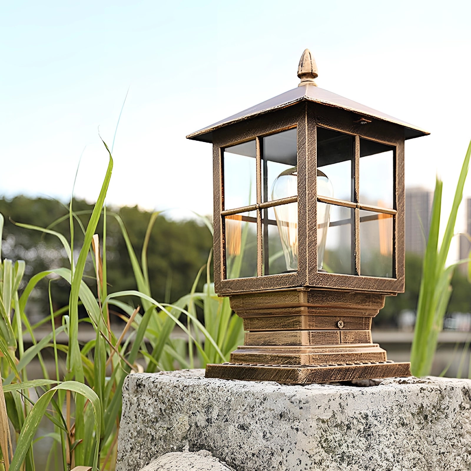 Classic vintage-style metal lantern placed outdoors, surrounded by greenery including grass, trees, and a wooden gazebo under a clear sky, creating a cozy, rustic landscape.