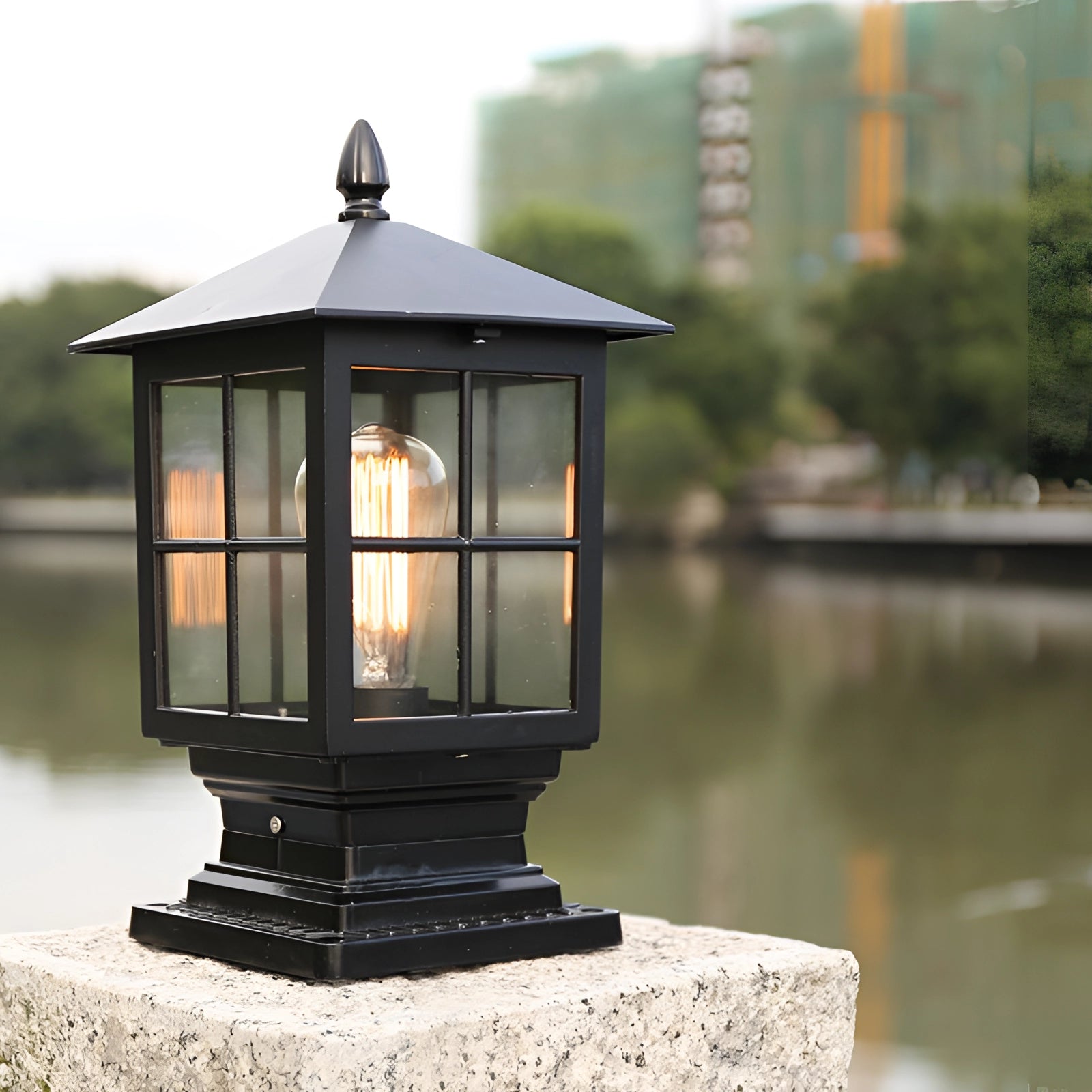 Classic vintage-style black metal lantern placed outdoors near a body of water, with trees and a clear sky reflected on the surface.