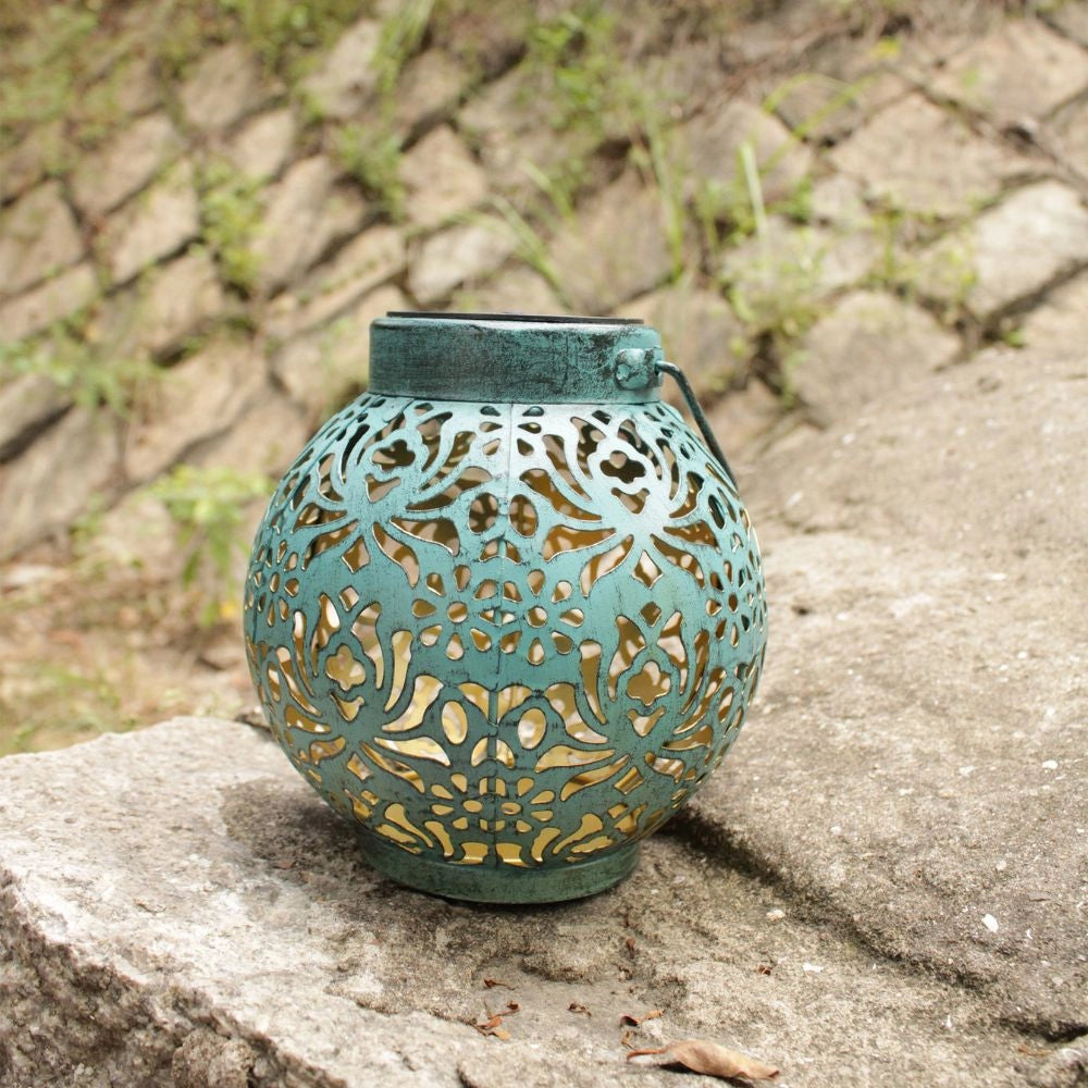 A decorative solar lantern with intricate floral cut-out patterns designed to project beautiful shadows, placed outdoors among plants and trees. The lantern has a pottery-like finish with shades of brown, resembling an ancient artifact.