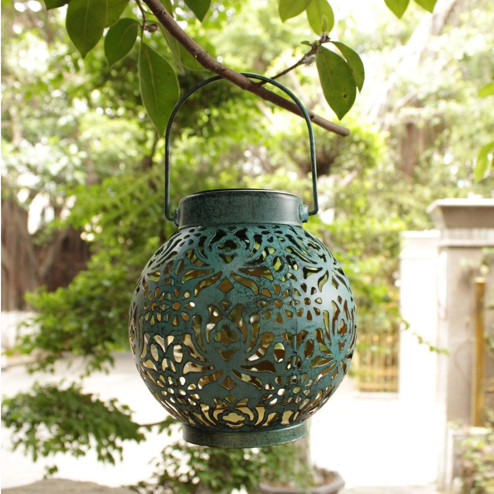 Decorative solar lantern with intricate floral cut-out designs, projecting elegant shadows, placed on a wooden surface surrounded by potted plants and flowers.