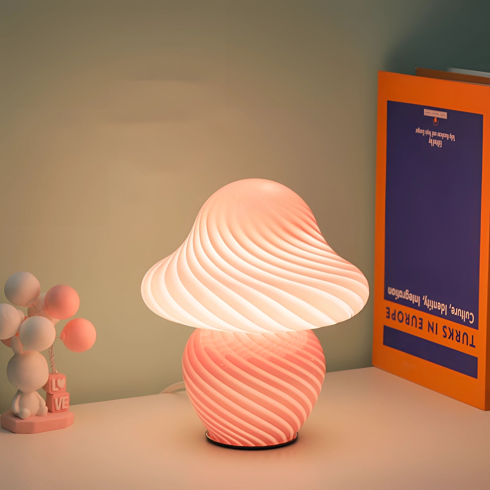 Elegant white spiral table lamp with a modern sculptural design, featuring a small pink swirl accent on the base. The lamp emits a soft orange light, creating a warm ambiance and artistic visual appeal. The circular pattern adds a unique touch to the contemporary space.