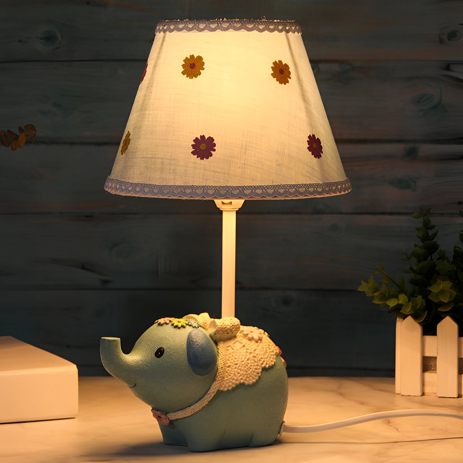 
A whimsical elephant table lamp designed for a nursery, featuring a decorative floral lampshade. The lamp is crafted with a wooden base and emits a soft glow, enhancing interior design with its artistic and playful aesthetic.