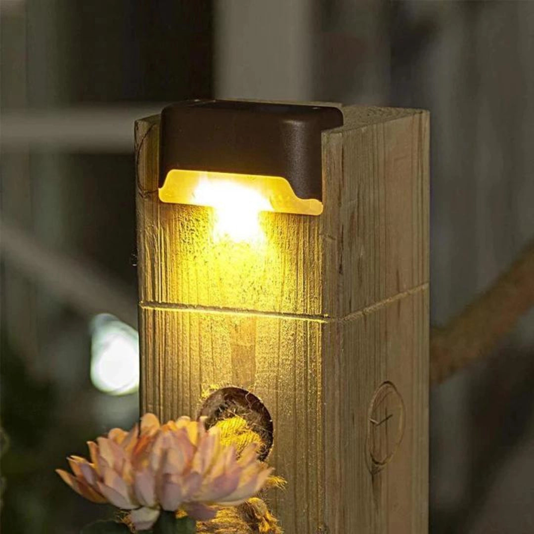 Energy-efficient LED waterproof deck lights illuminating an outdoor patio area, surrounded by wooden deck flooring and garden plants with flowers. A modern light fixture with a cylindrical metal design is visible, casting a warm glow on the surroundings.