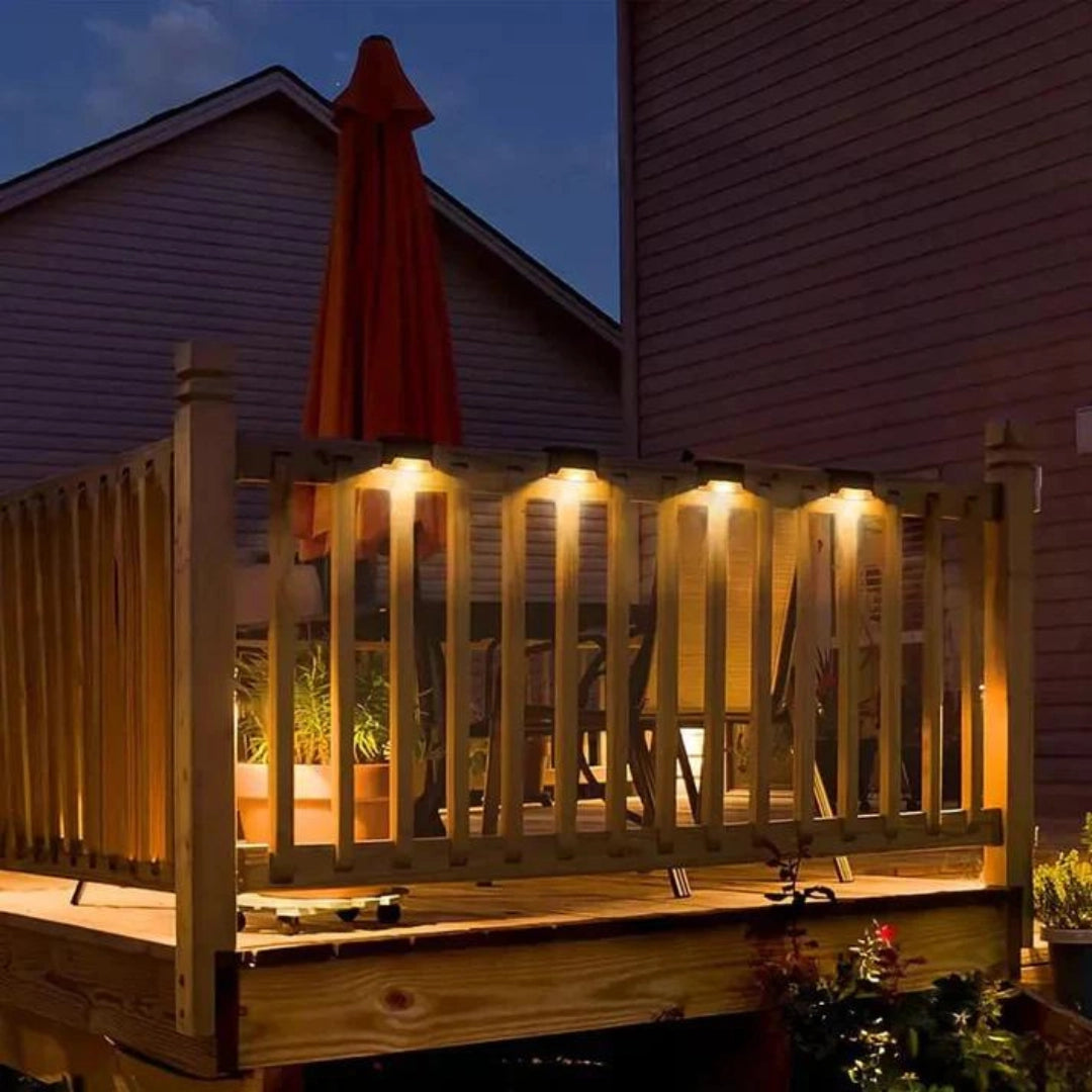 Energy-efficient LED waterproof deck lights installed on an outdoor patio, illuminating wooden surfaces and surrounding landscape, with a backdrop of sky and building facade, accompanied by flowerpots and a fence.