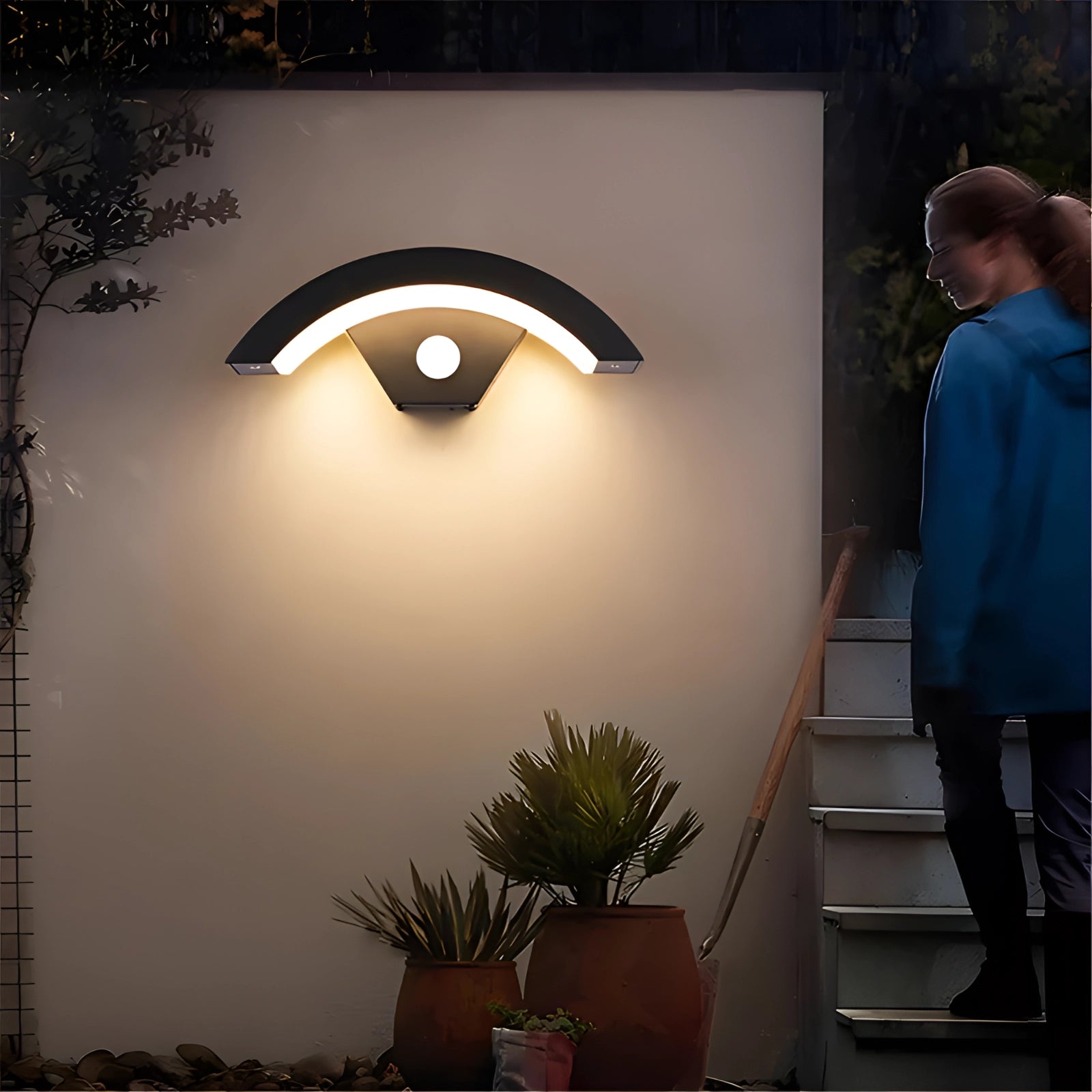 Eye-shaped LED wall light with a modern design mounted on an exterior wall, surrounded by lush green plants and wood accents, creating an inviting outdoor ambiance.