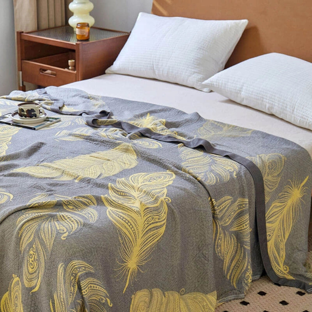 Floral yellow leaf pattern reversible blanket displayed on a bed with grey bed linen decor, complemented by pillows and surrounded by a cozy interior design setting featuring a window and wooden furniture elements.
