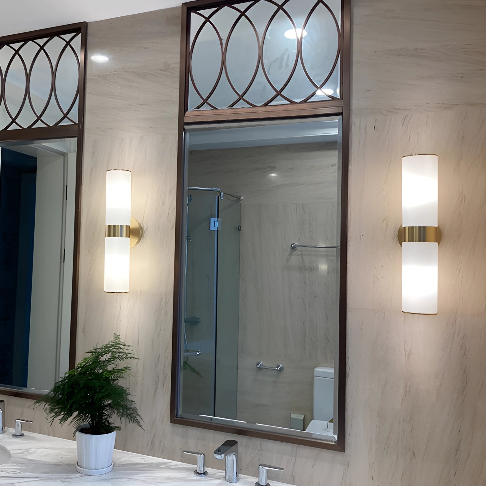 Gold bathroom sconce with a cylindrical frosted glass design mounted on a wall in a stylish interior setting, with a touch of greenery visible.