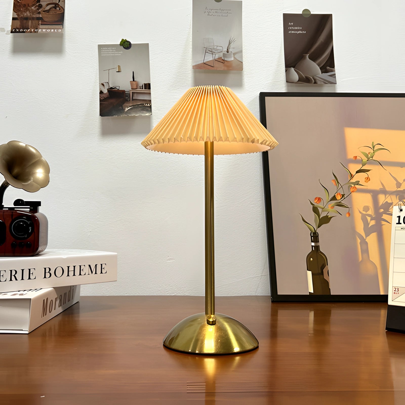 A gold brass floor lamp with a pleated shade in a vintage mid-century retro style, positioned next to a wooden table with shelving and a potted plant in the background.