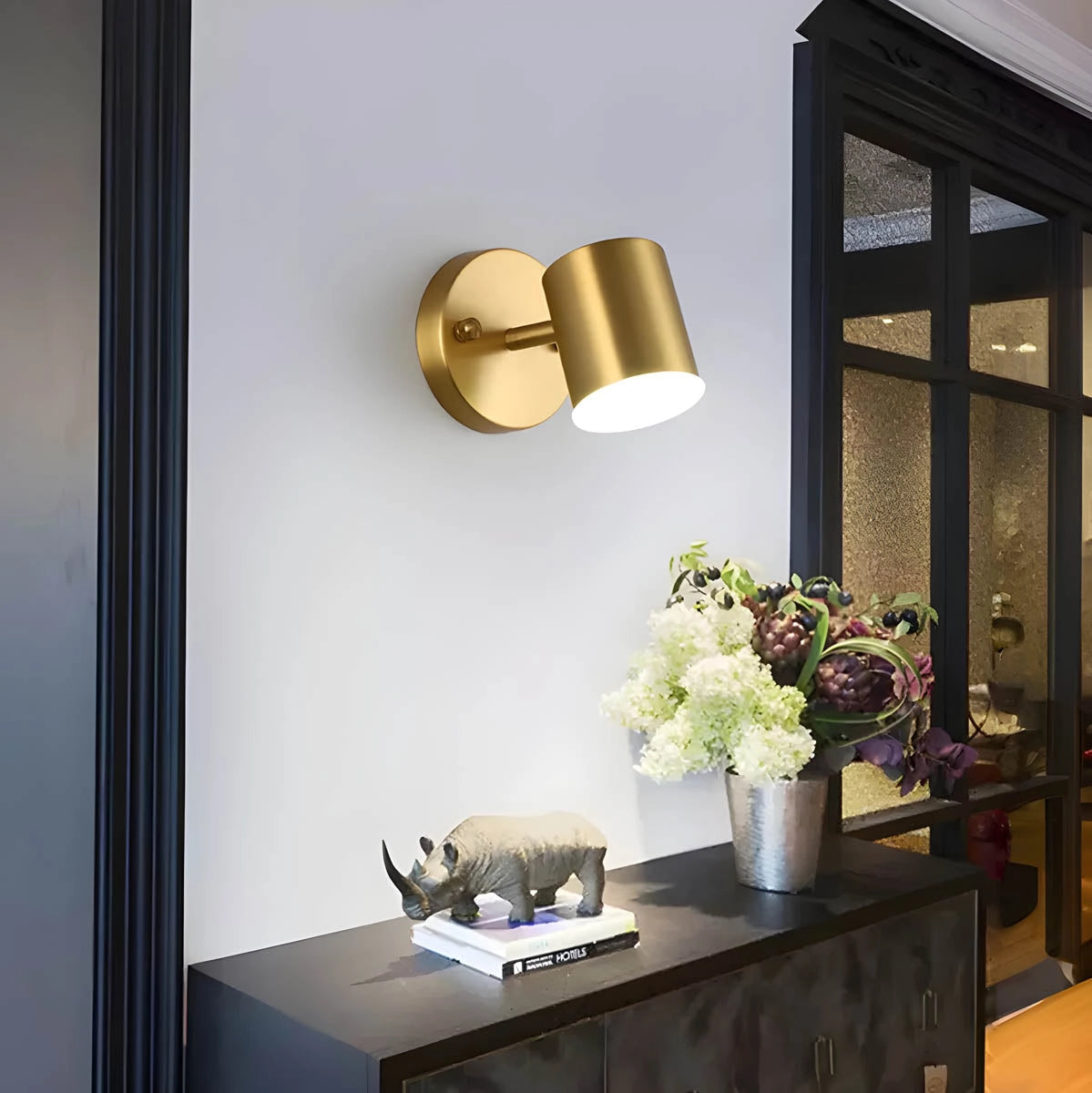 Gold LED wall lamp with a modern brass finish, mounted on a wall next to a window, casting light on a decorative interior setting with a table adorned with a vase containing flowers and a nearby houseplant.