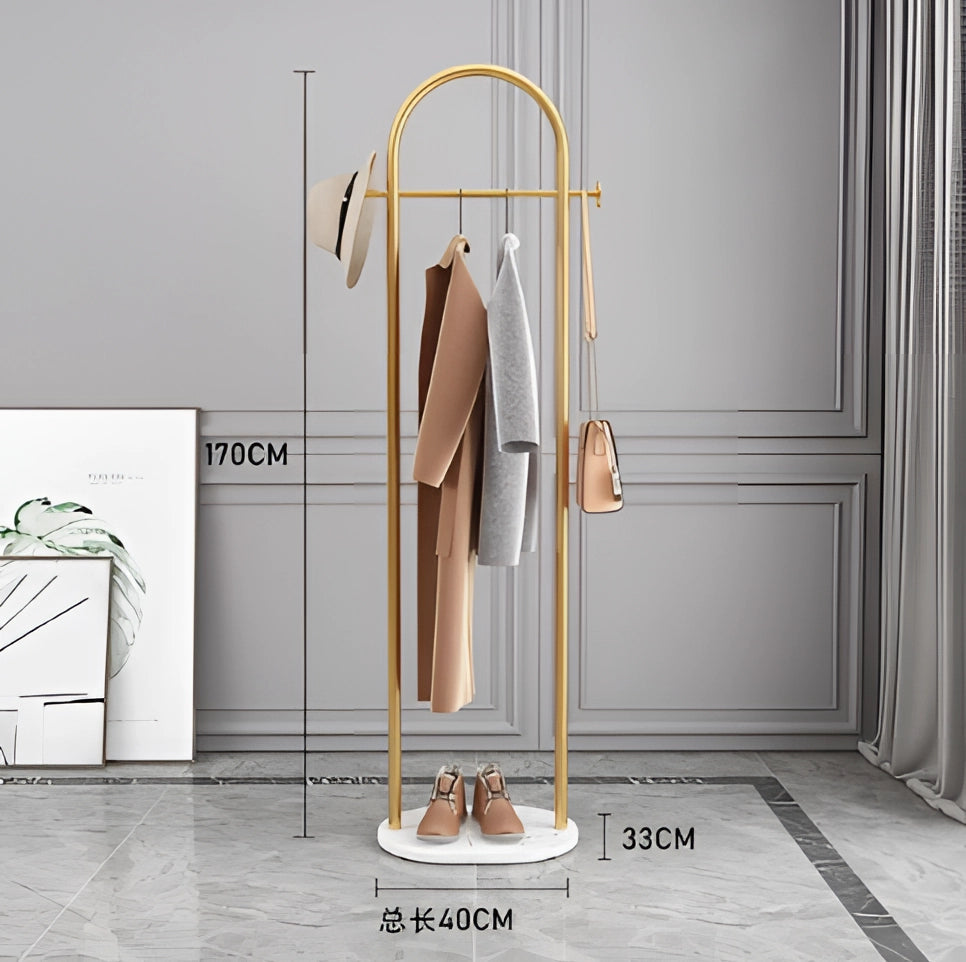 Gold metal coat rack with a sturdy marble base, featuring a minimalist and luxurious design, suitable for hanging garments. The coat rack is positioned on a wooden floor near a door, showcasing its elegant functionality and fashion-forward style.