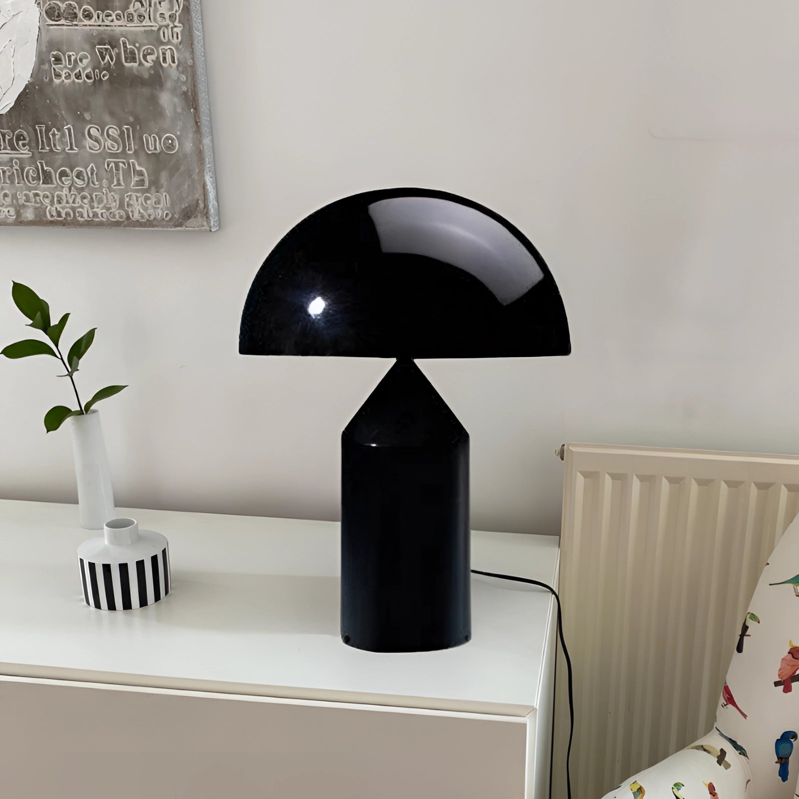 Gold mushroom table lamp with luxury brass finish, displayed on a black table in a modern bedroom setting, featuring a sleek design alongside white and grey decor elements, including plants and wooden shelving.