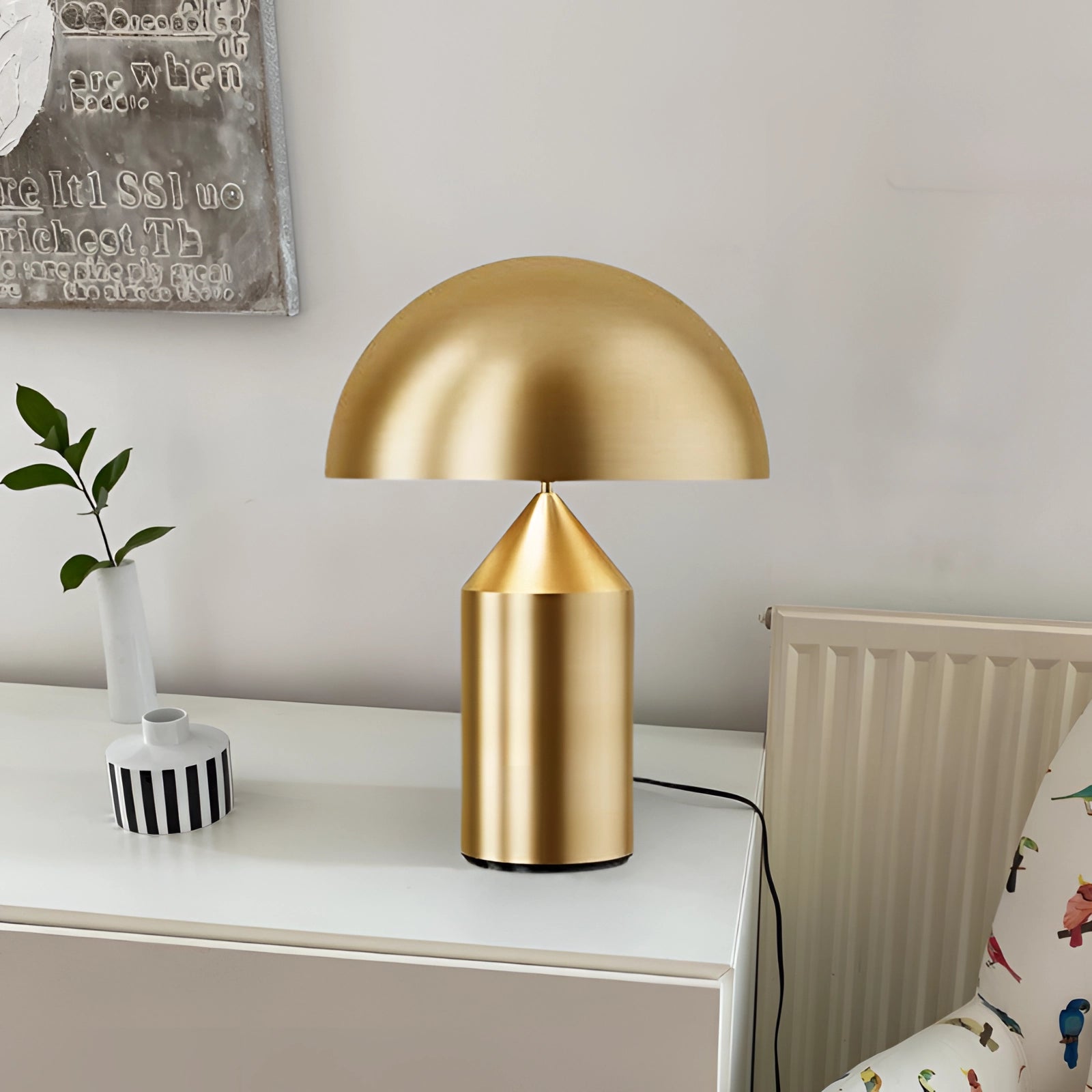 Gold mushroom table lamp with a luxurious brass finish displayed on a rectangular wooden table, complementing modern bedroom interior design. The lamp is surrounded by decorative plants and shelving, enhancing the elegant and stylish ambiance.