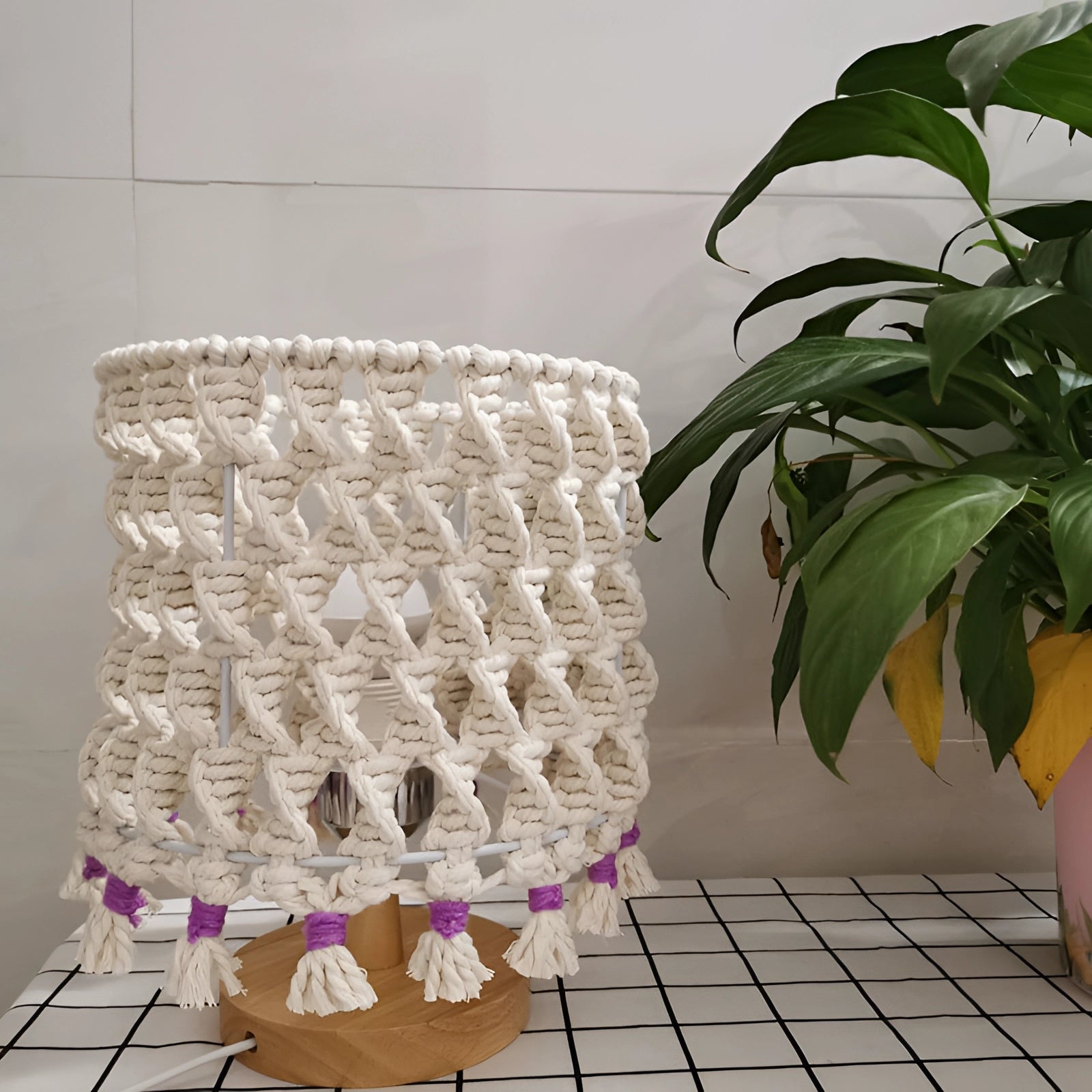Handmade boho macrame lampshade with intricate tassel design, featuring natural materials, displayed in a room with a wooden floor and potted houseplants.
