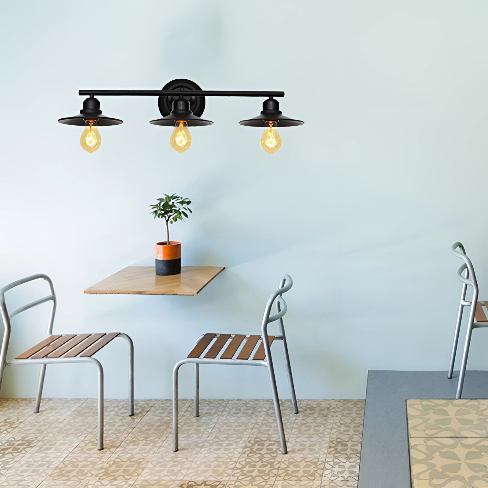 Industrial geometric metal cage pendant light hanging from the ceiling, illuminated by an Edison bulb, in a stylishly decorated interior setting with a wooden table, chairs, houseplant in a flowerpot, and decorative shelving on the wall.