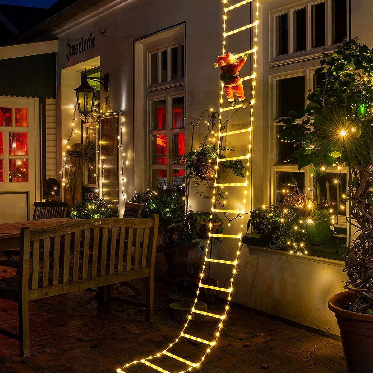 Santa Claus figure climbing on a building facade, holding a festive garland with LED lights, surrounded by potted plants and window decorations, creating a cozy holiday scene.