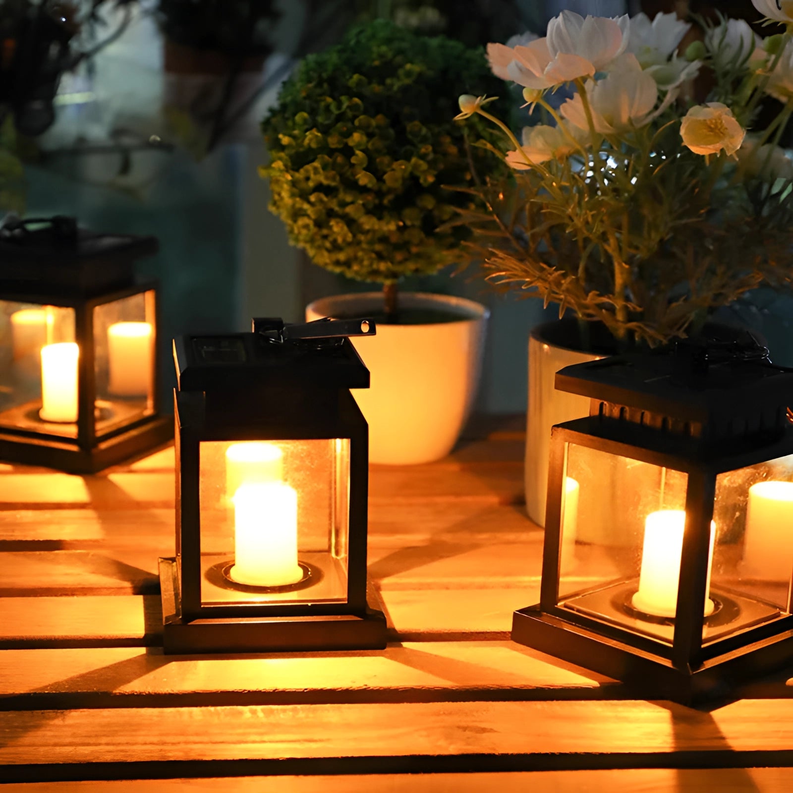 LED decorative outdoor lanterns made of black metal, featuring a waterproof design with a flameless lighting element, placed on a table surrounded by plants and flowers, casting a warm yellow glow.