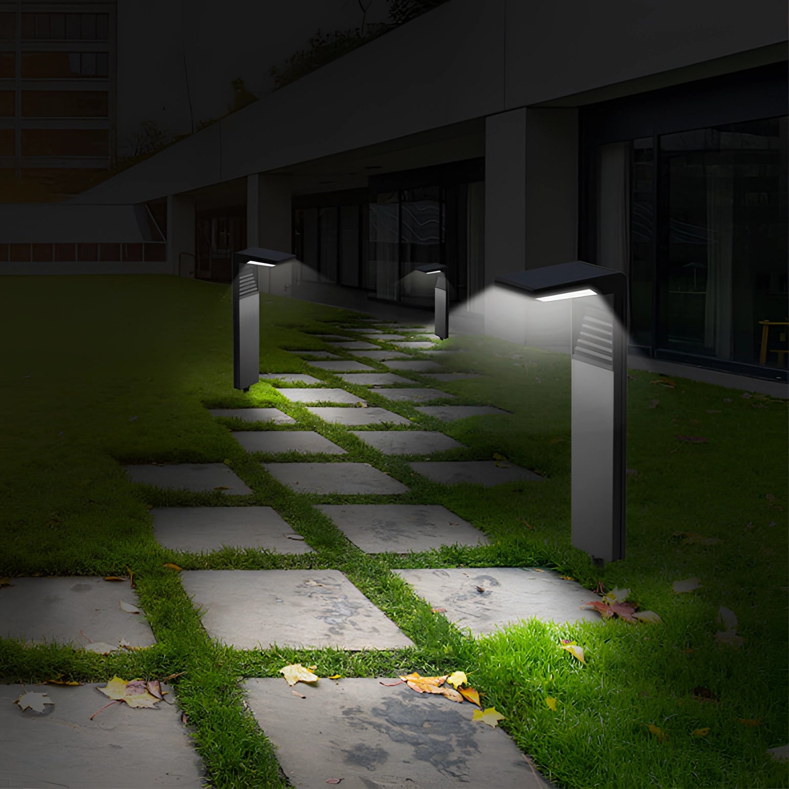 LED solar pathway lights installed along a sidewalk, surrounded by grass and plants, with a building wall visible in the background.