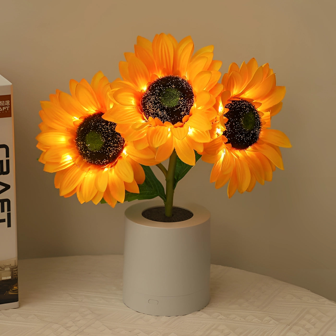 LED sunflower lamp with bright orange petals and a green stem, displayed in a white flowerpot. The lamp resembles a real sunflower and is designed as a decorative night light for home interiors.