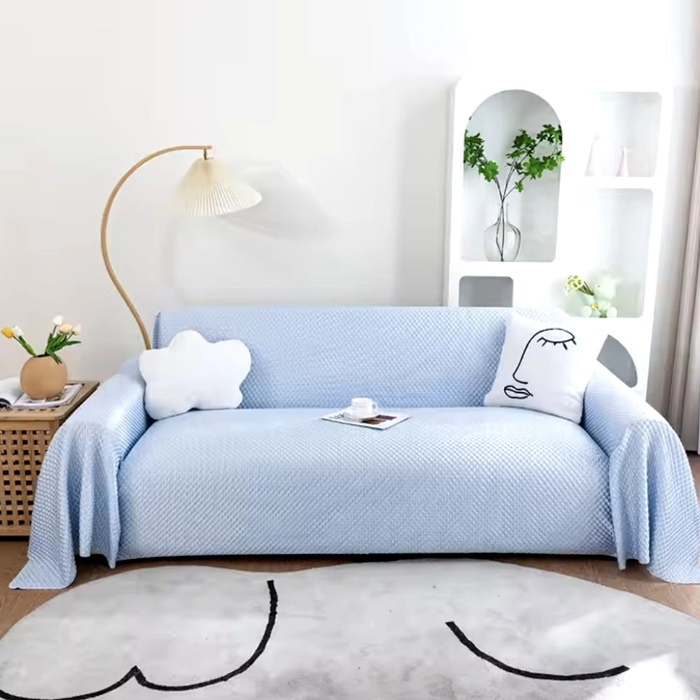 Light blue quilted anti-slip sofa cover in a cozy living room setting, featuring a plant, white and grey decor, wooden floor, and a pillow, measuring 210x300cm, designed to be pet-friendly and washable.