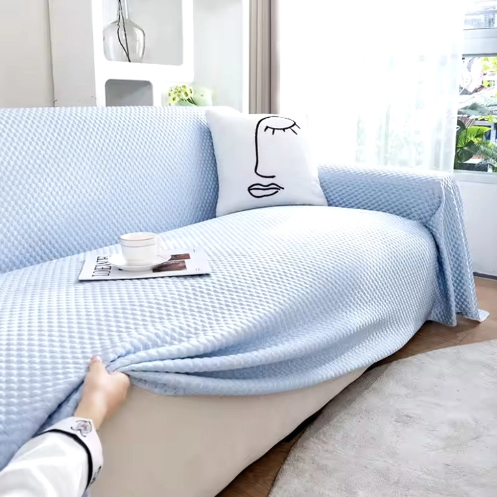 Light blue quilted anti-slip sofa cover draped over a rectangular sofa, set in a cozy interior with a wooden floor and grey accents. The cover is pet-friendly and washable, enhancing comfort and protection for furniture. A pillow is placed atop the sofa, and a window is visible in the background.
