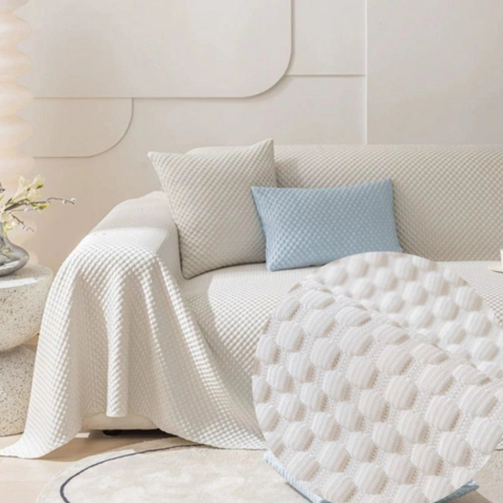 Light blue quilted anti-slip sofa cover displayed on a beige studio couch in an interior setting, featuring a comfort-enhancing design suitable for pets, with a white pillow and a nearby plant, offering a cozy and stylish addition to home decor.