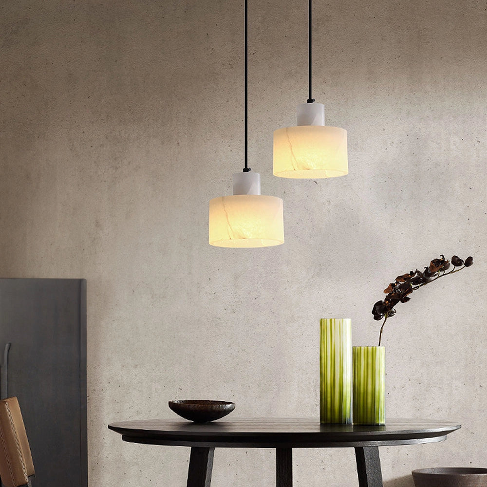 A luxury alabaster pendant light with a natural stone finish, hanging from a minimalist ceiling in a contemporary indoor setting with modern furniture and plants.