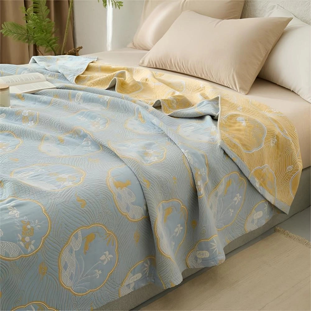 Luxury blue bedding set featuring an ornate gold floral pattern, displayed on a wooden bed frame with matching bed skirt, in a well-lit bedroom with hardwood flooring and a large window.