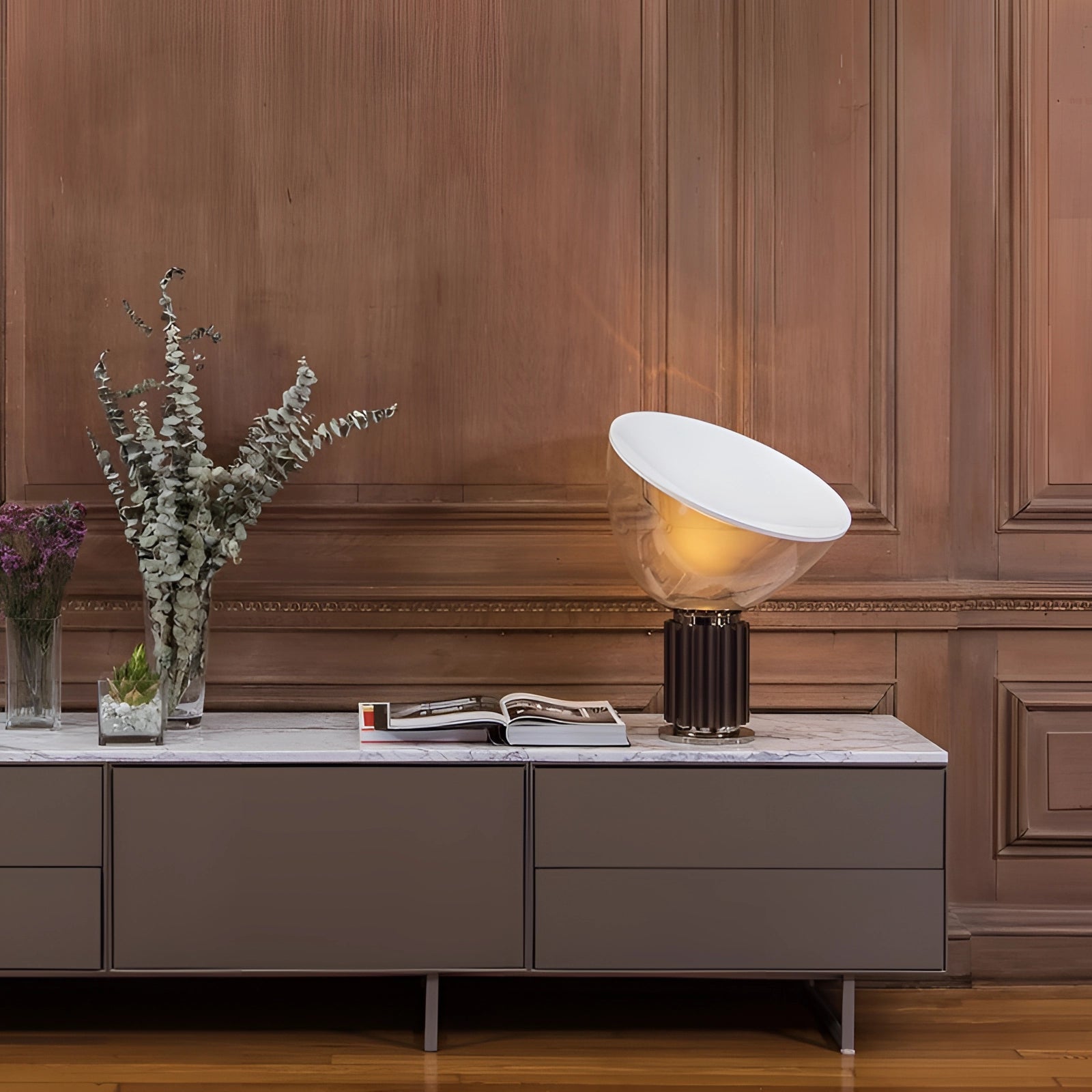 Luxury designer glass lamp with a modern ribbed base and dome placed on a wooden table in a stylish interior setting with a cabinet and plant in the background.