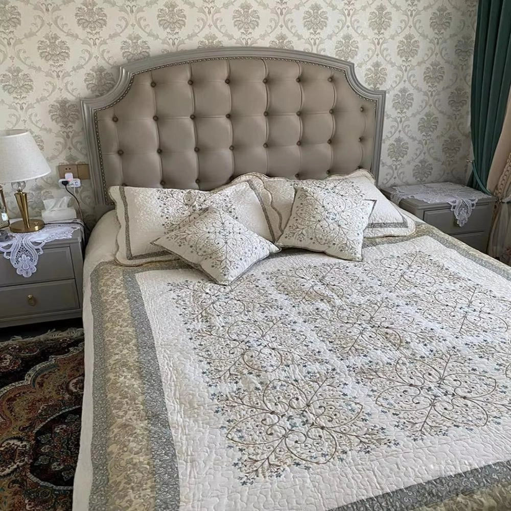 Luxury floral quilt set displayed on a neatly made bed with embroidered pillowcases and scalloped edges, set in a comfortable and elegantly furnished room with beige and grey tones.