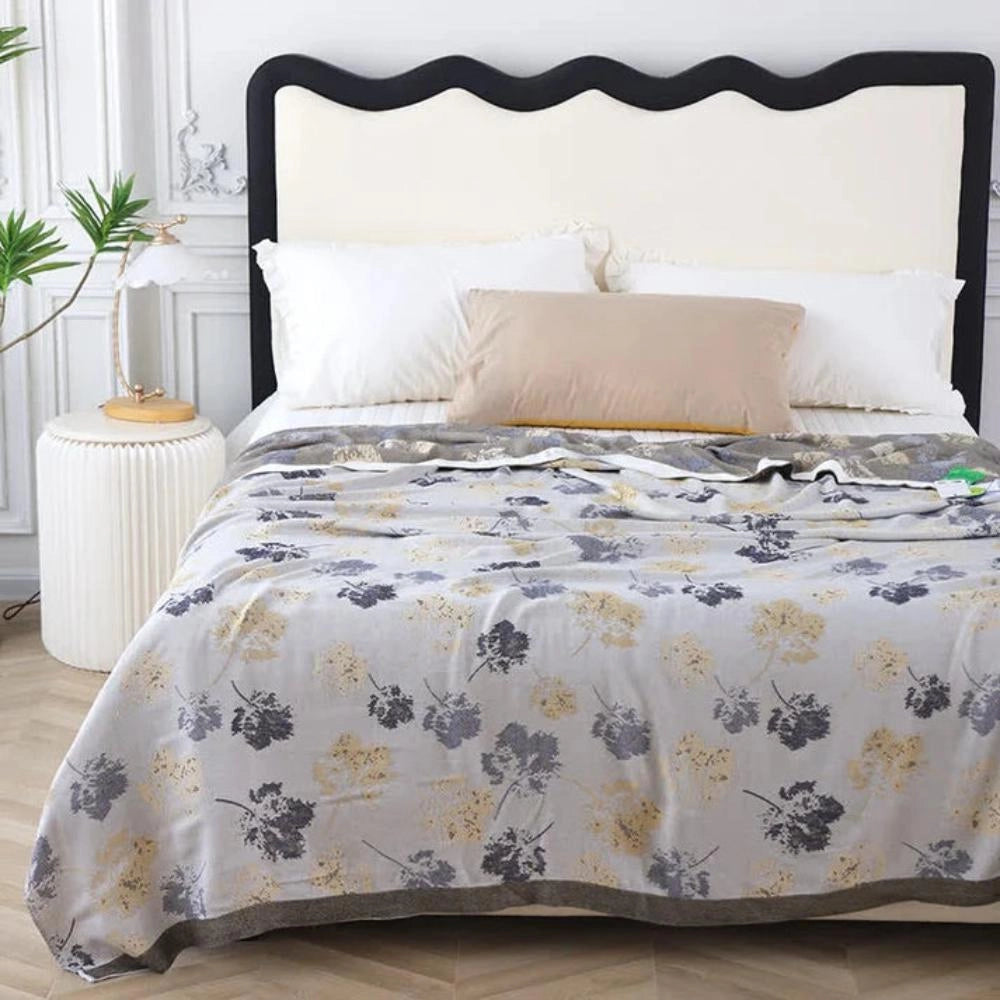 Luxury floral silk comforter in grey brown draped over a bed with a wooden frame, surrounded by elegant interior decor including houseplants and furniture, creating a cozy and stylish bedroom setting.