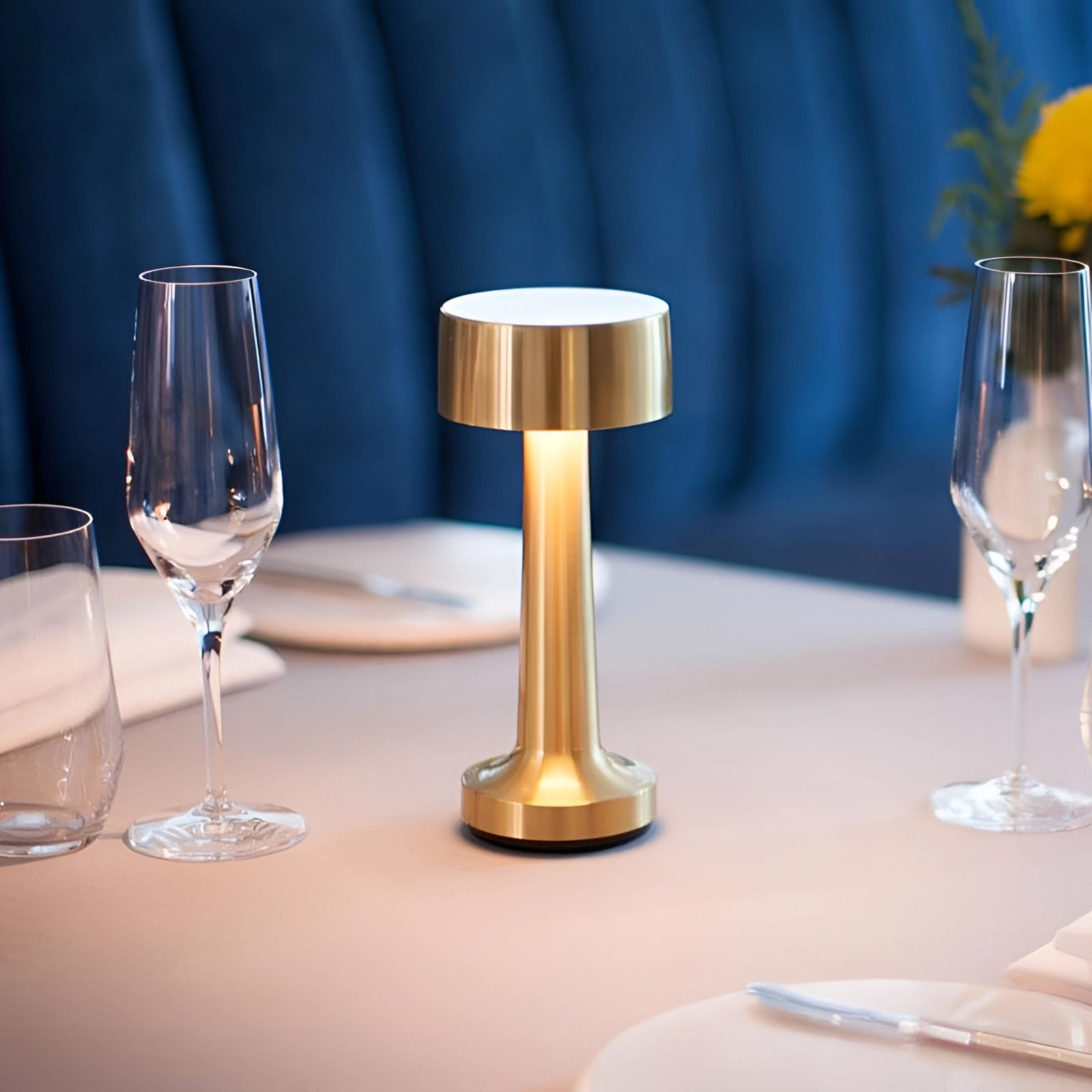 Luxury gold cordless LED table lamp with a sleek design, set on a wooden surface next to elegant glassware, including a wine glass and champagne stemware, with a warm yellow light illuminating the scene.