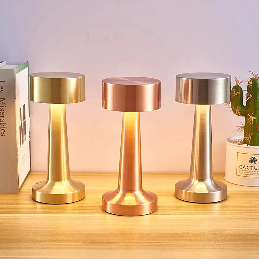 A luxury gold cordless LED table lamp with a sleek cylindrical design, placed on a wooden surface next to a wine glass.
