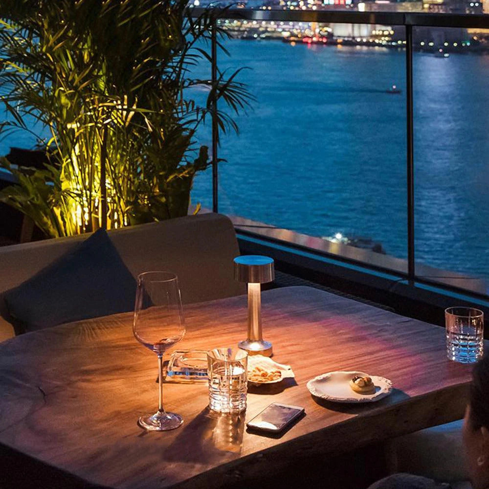 Luxurious gold cordless LED table lamp placed on an elegantly set table, accompanied by tableware and a wine glass, with an azure background suggesting a sophisticated indoor or outdoor setting.