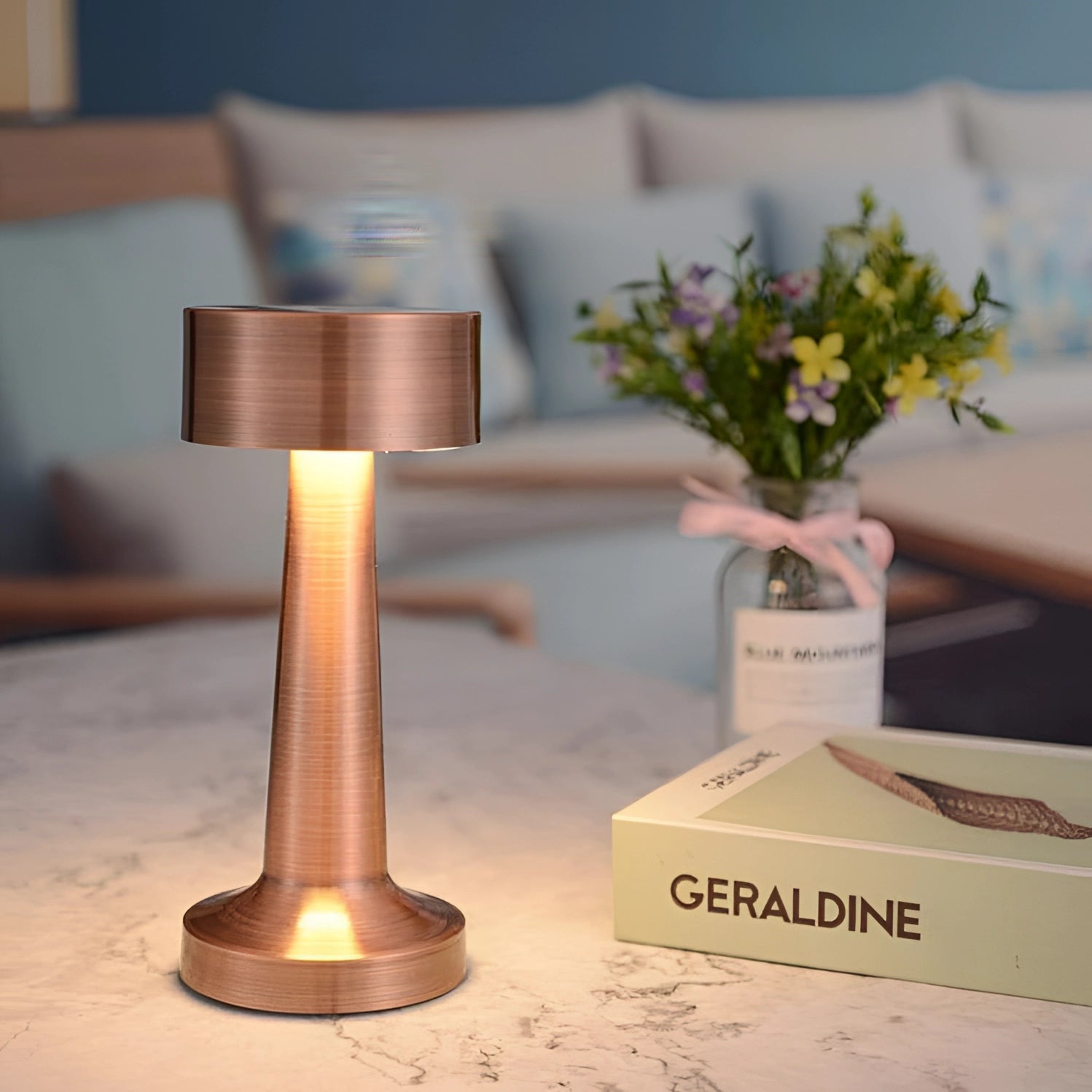 Luxury gold cordless LED table lamp on a wooden table, surrounded by white flowers and green plants, creating a decorative and elegant setting.