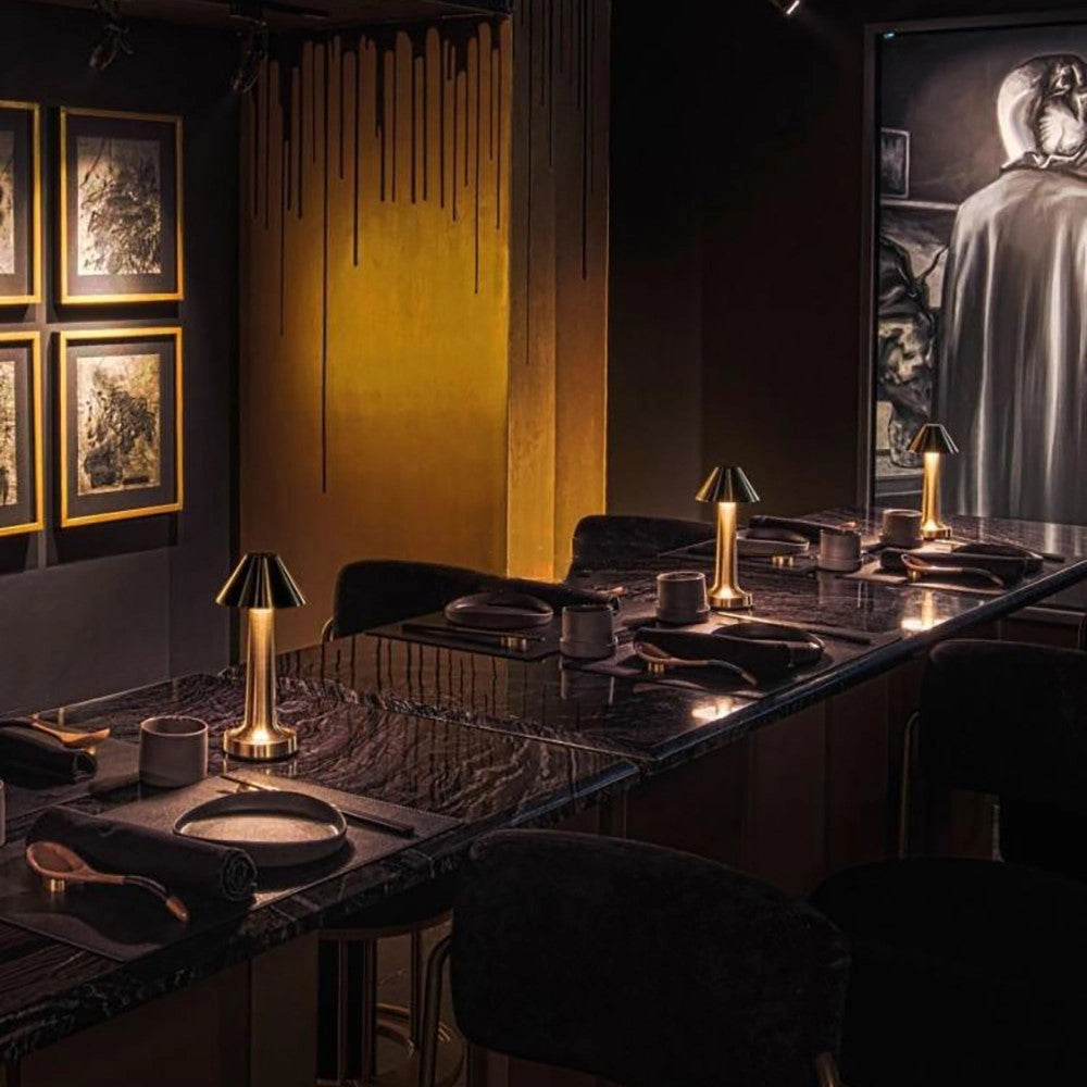 A luxurious gold cordless table lamp with a rechargeable battery, elegantly placed on a dining table with tableware, set in a stylish black and wood interior design setting.