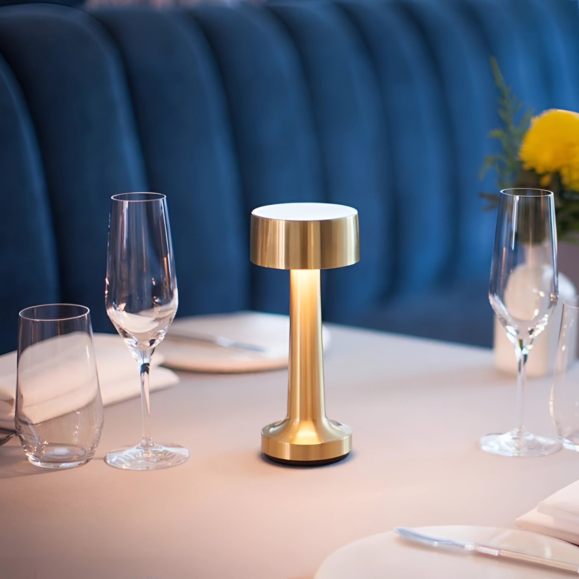 Elegant rechargeable gold cordless table lamp with a cylinder shape, placed on a dining table set with tableware, wine glasses, and a tablecloth.