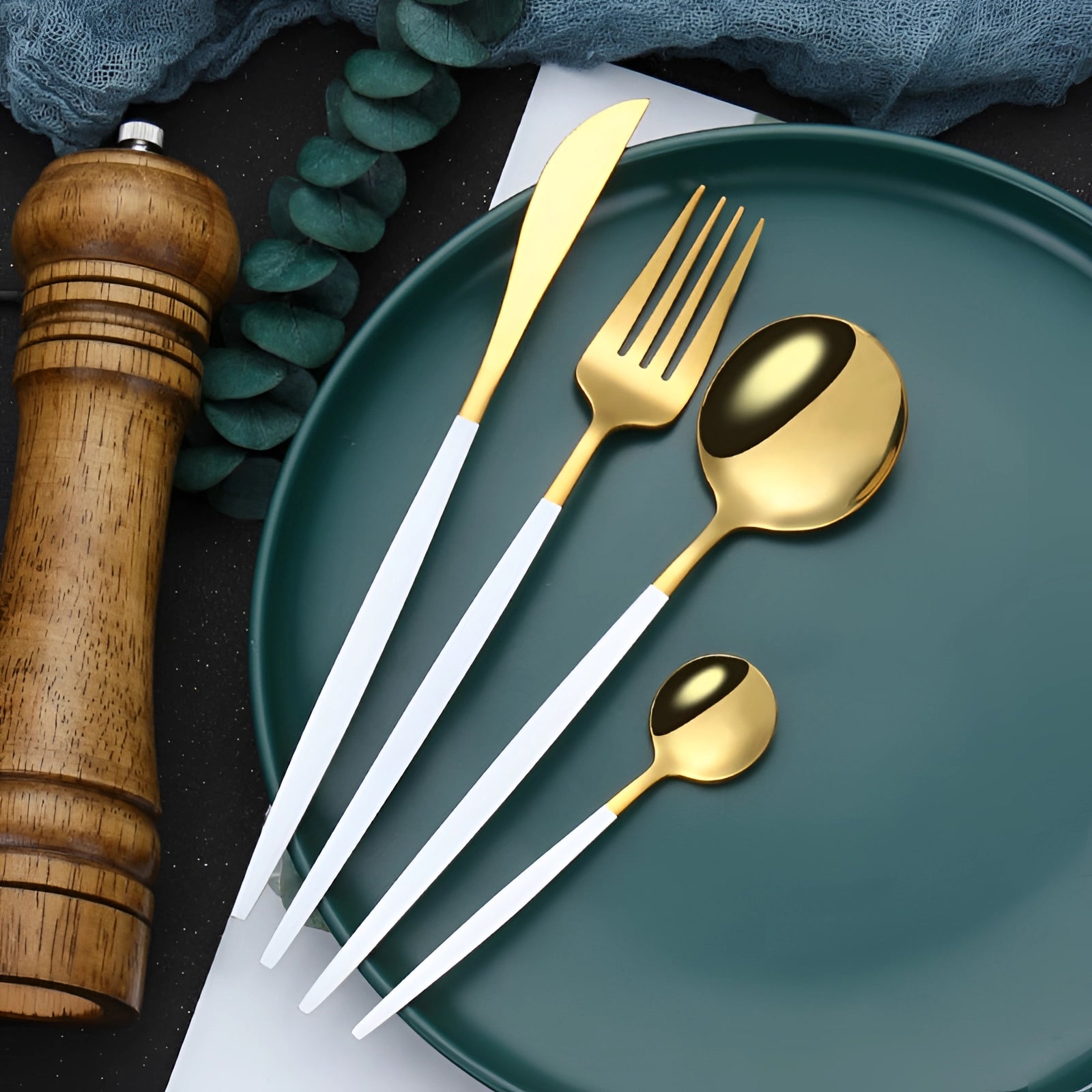 Elegant gold cutlery set displayed on a white table setting, featuring a gold fork, knife, and spoon placed on a light wood table surface with a white plate and tablecloth.