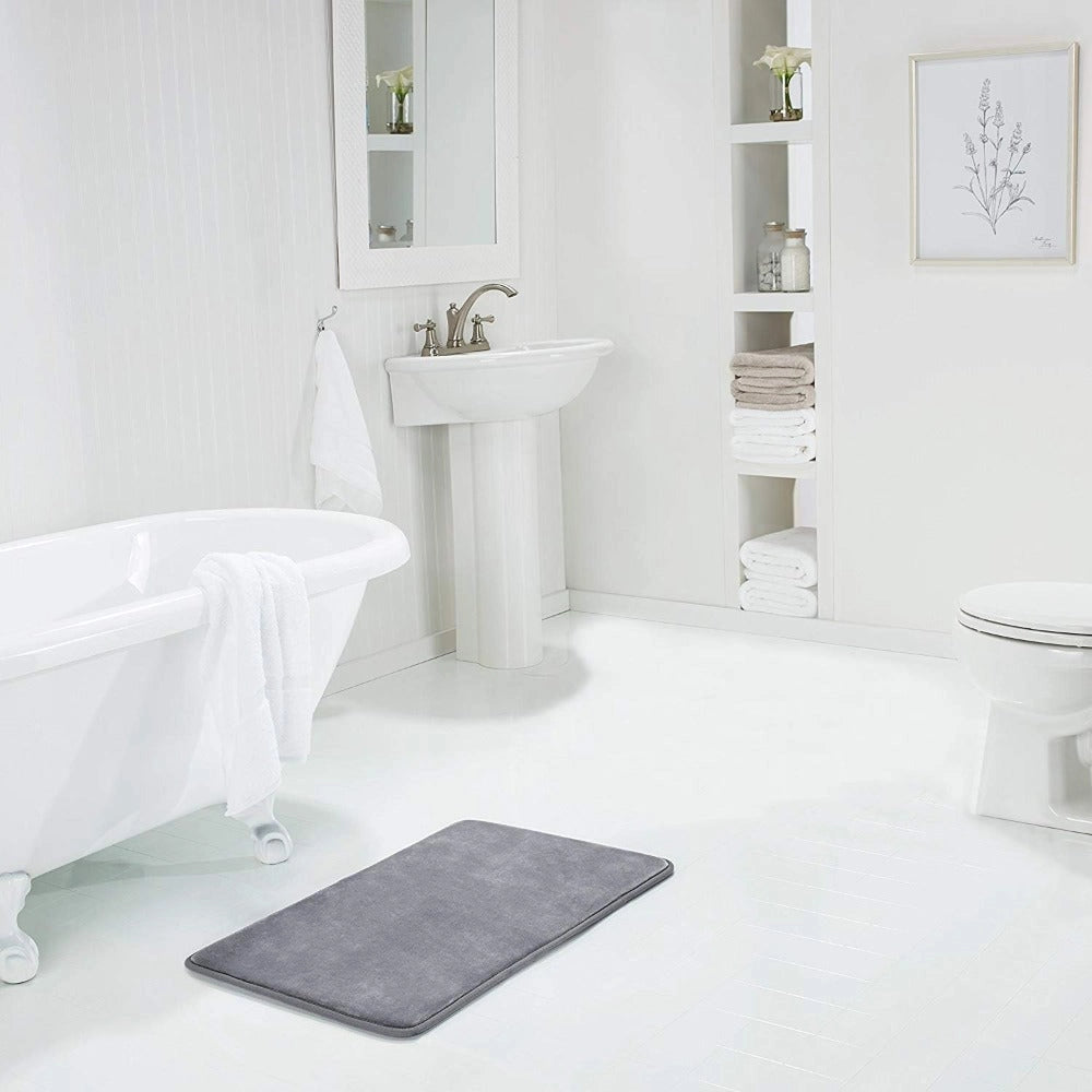 Luxurious grey memory foam bath mat with a plush surface, designed in a rectangular shape. It is placed in a contemporary bathroom setting, complementing the sink and bathtub fixtures. The mat is non-slip and quick-drying, enhancing the bathroom's elegant and functional design.