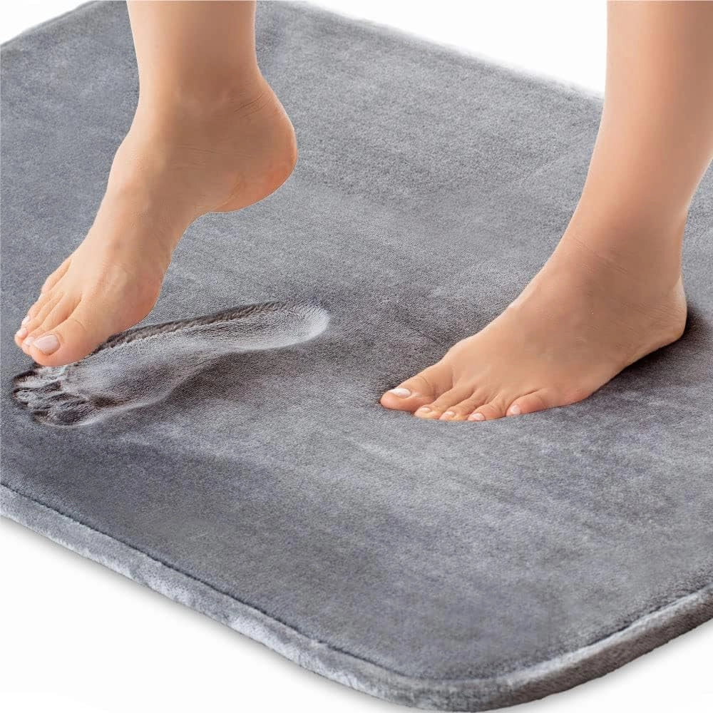 Luxury grey memory foam bath mat featuring a non-slip backing, quick-dry functionality, and plush texture, shown in an extra-large size (60x90cm) on a bathroom floor with a barefoot person standing on it.