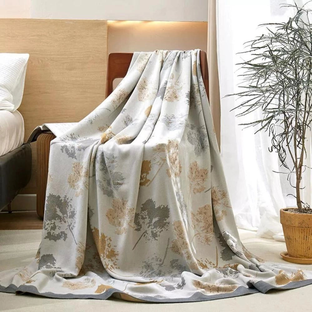 Luxury silk floral throw blanket in light tan and grey draped over a wooden chair in a stylishly decorated room with plants, wooden flooring, and large windows, enhancing the elegant home decor.