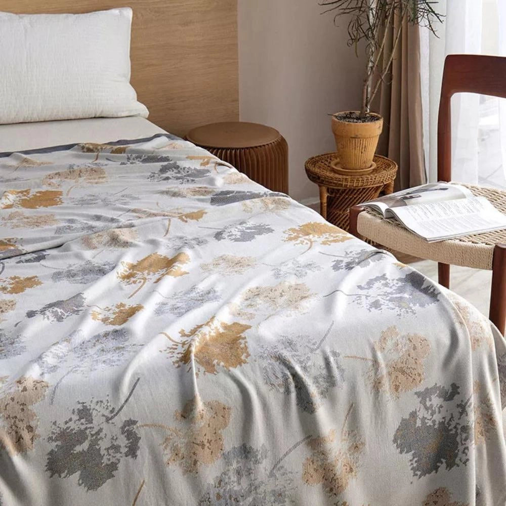 Luxury silk floral throw blanket draped over a modern wood bed frame in an elegantly designed bedroom, with white and neutral tones, featuring a plant and stylish flooring for a comfortable and sophisticated home decor setting.