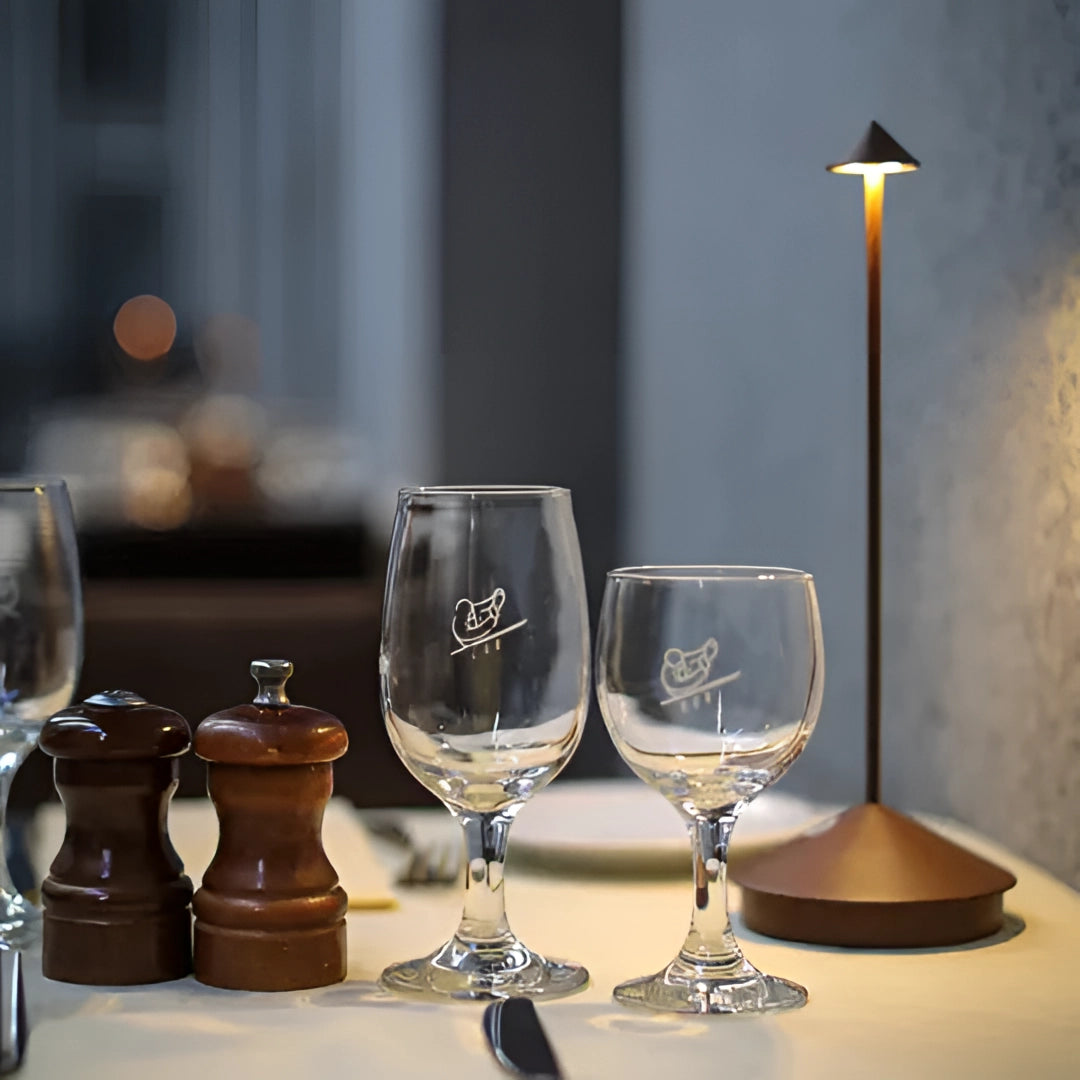 Minimalist arrow-shaped LED desk lamp with a sleek design, positioned on an elegantly set table with various types of drinkware, including wine glasses and champagne flutes, showcasing a modern decor theme.