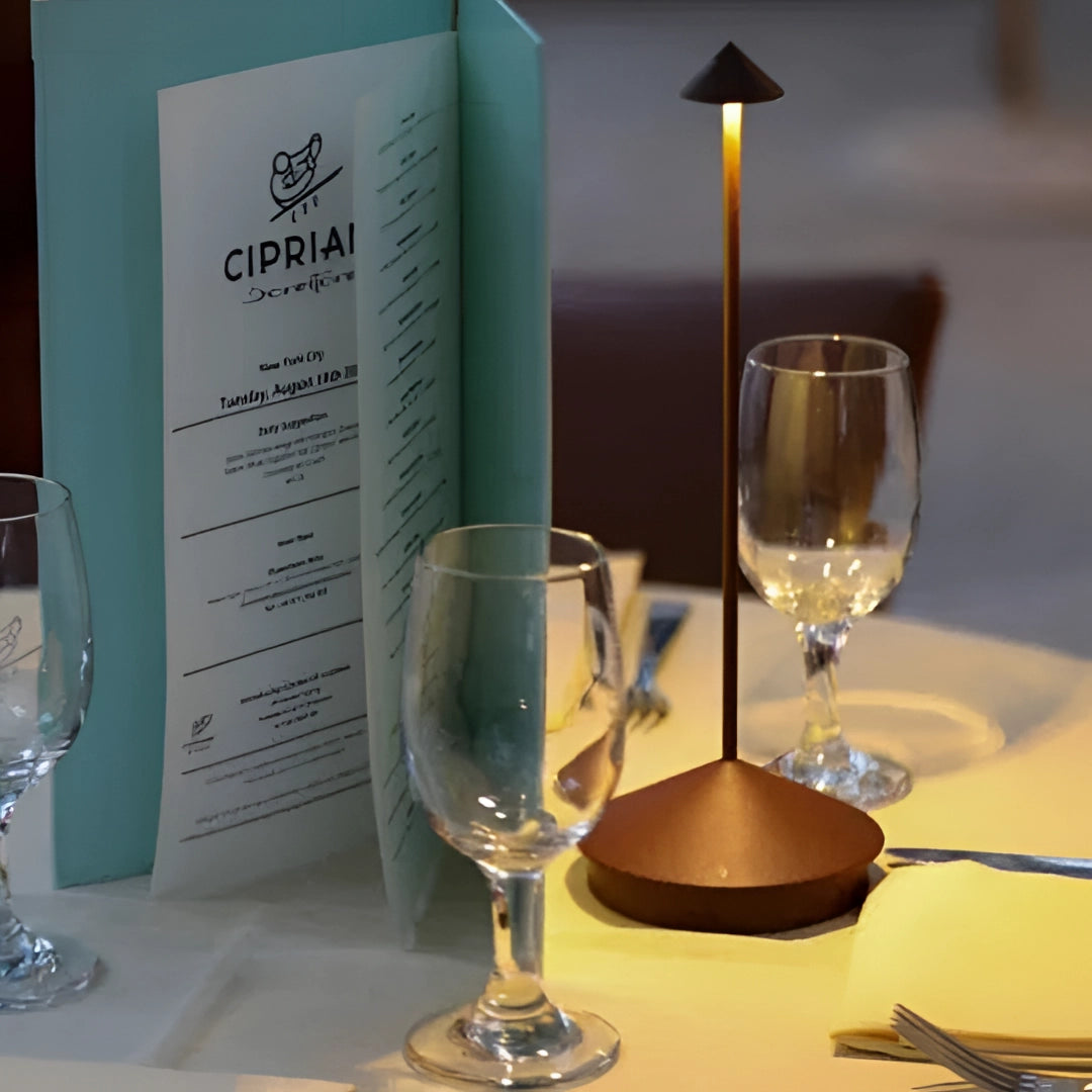 Minimalist arrow-shaped LED desk lamp with a sleek modern design, placed on a table surrounded by various drinkware including glasses and a bottle, adding a contemporary and stylish touch to the decor.