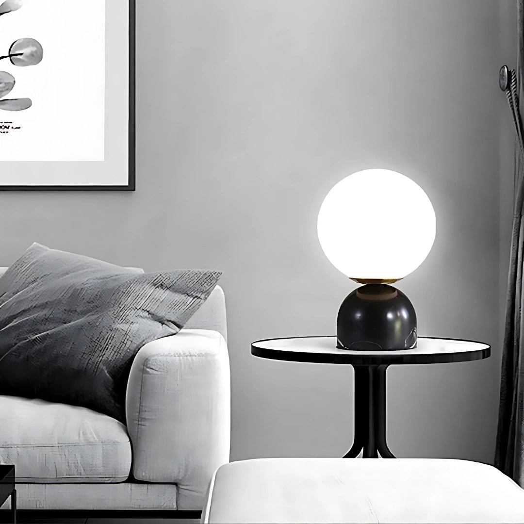 Minimalist black marble table lamp with a modern orb globe design, styled in Scandinavian decor, positioned on a white surface with a nearby couch in the background.