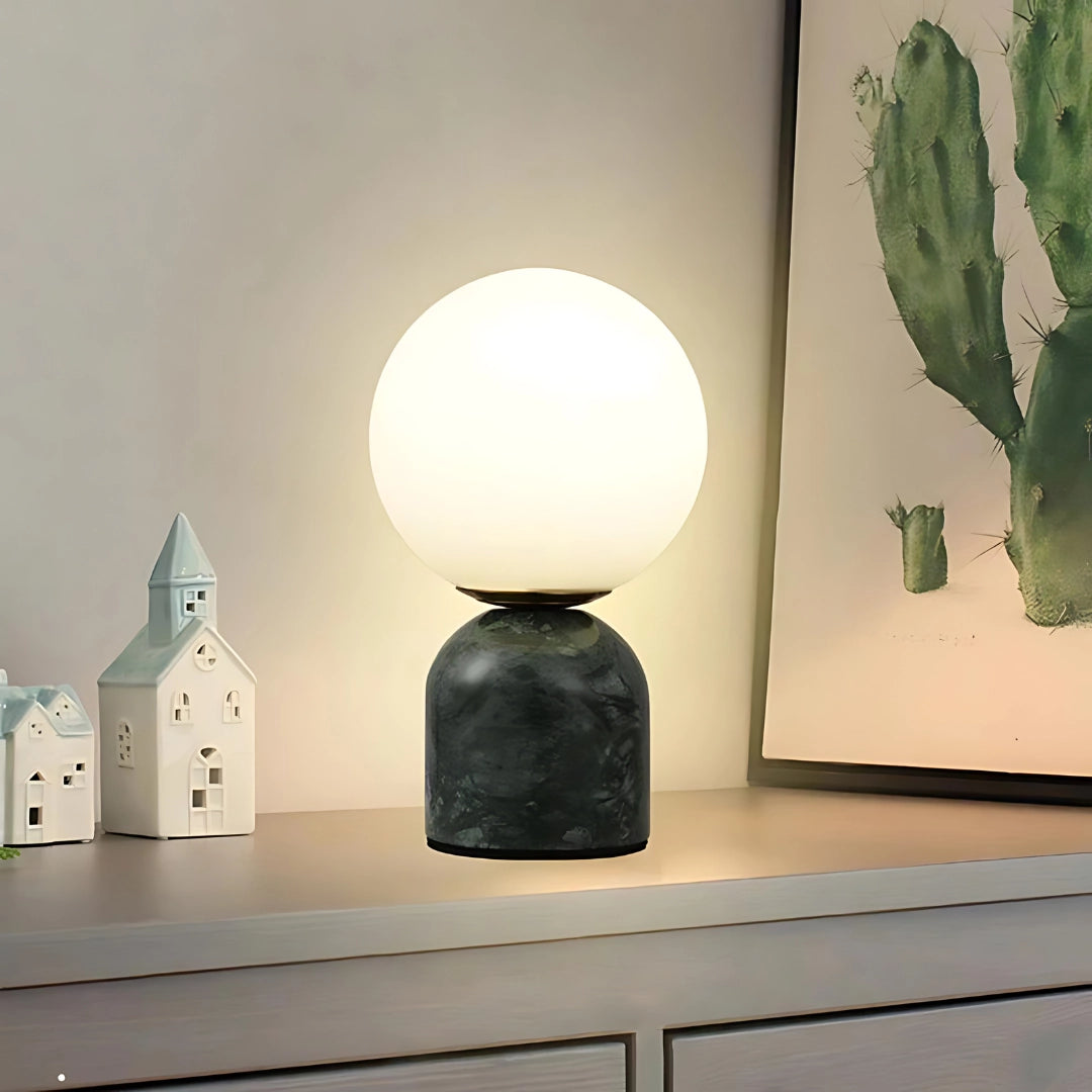 Minimalist marble table lamp with a dark green orb globe displayed on a wooden surface, surrounded by Scandinavian decor elements including a potted plant and art pieces.